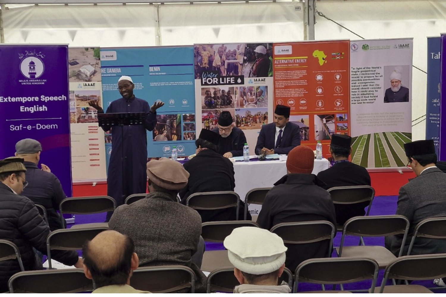 An extempore speech competition at the 2025 Ijtema — a competitor speaking from a lectern to a seated audience, with judges at a table and the competition banners behind