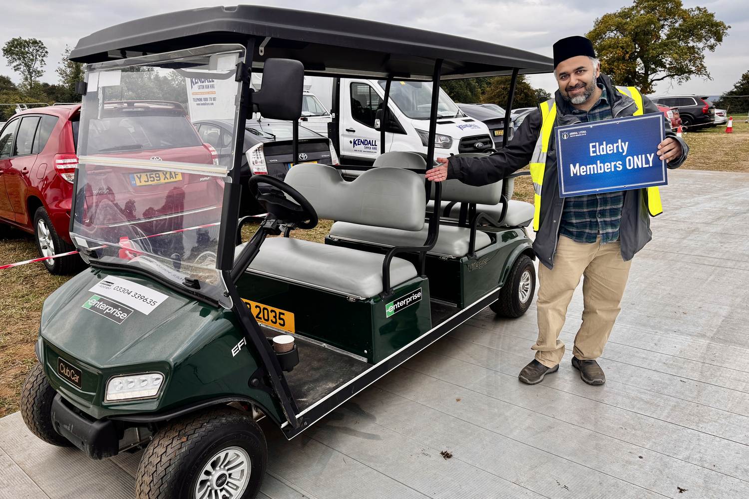 A volunteer in a high-visibility vest standing beside an electric buggy at the 2025 Ijtema, holding a sign reading Elderly Members ONLY