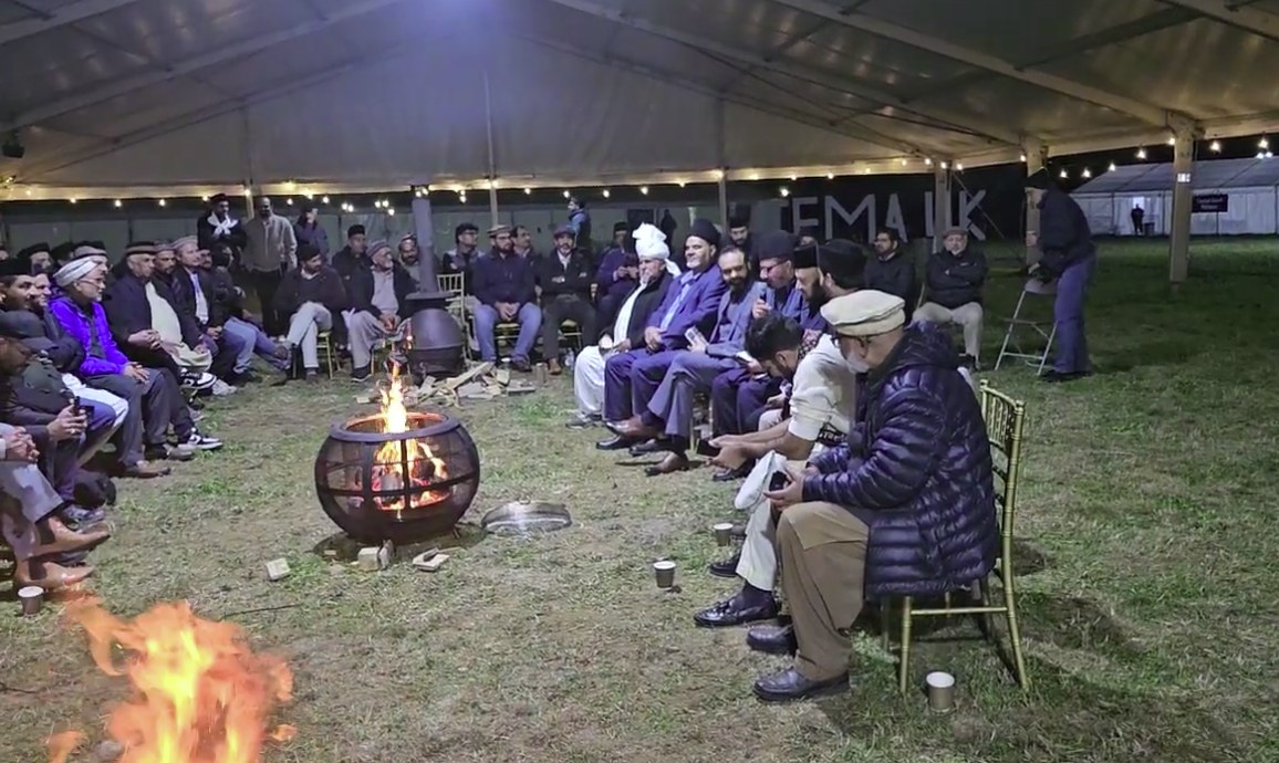 The Mushairah at the 2025 Ijtema after dark — men seated in a wide circle under a marquee canopy strung with lights, braziers burning between them and one of the reciters holding a microphone