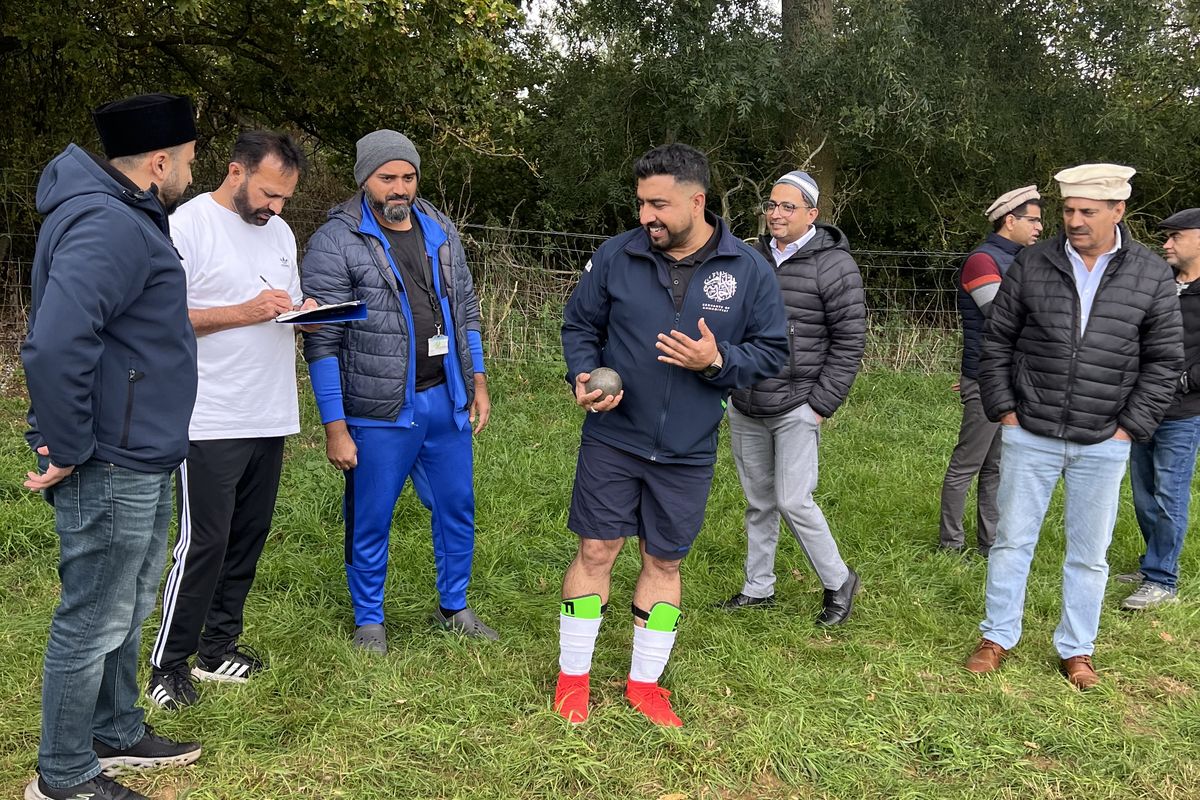 Members gathered on the grass at the 2025 Ijtema watching a shot put attempt, one of them recording scores on a clipboard