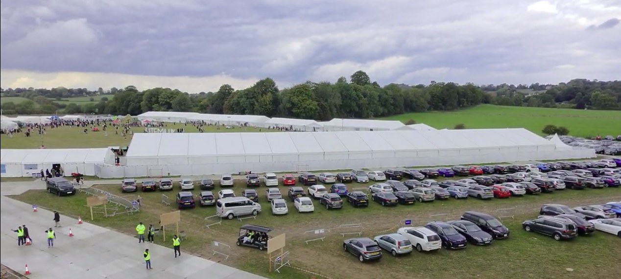The Ijtema site at Puttenham seen from above in daylight, the long marquees running across the field with the visitor car park filled alongside them and marshals directing arrivals