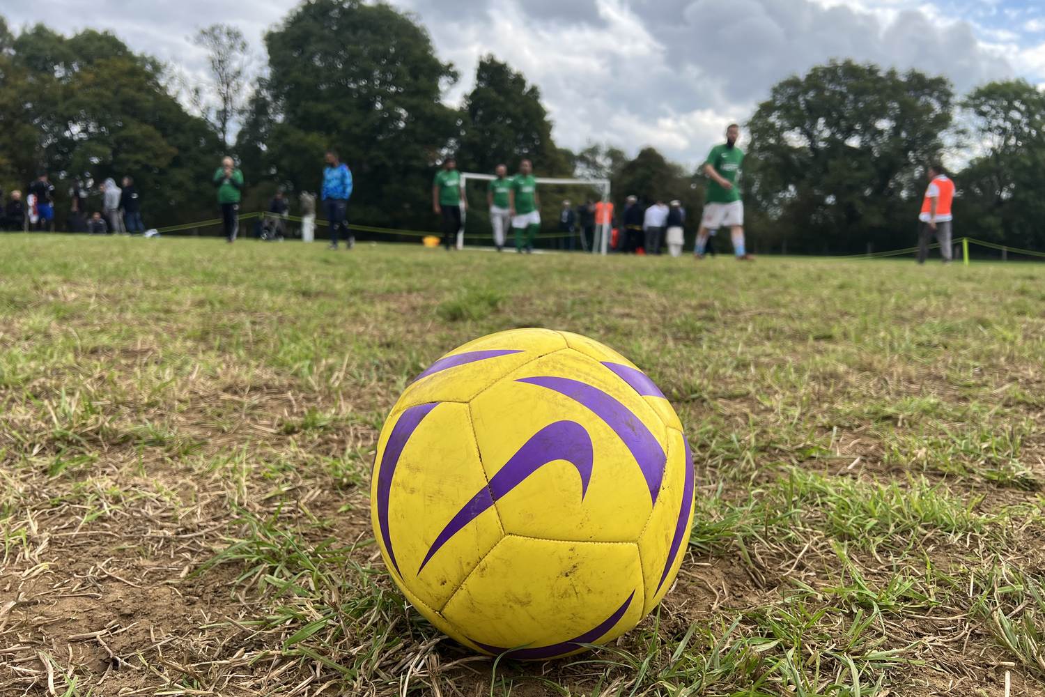 A football resting on the grass at the 2025 Ijtema, with players in kit and a goal on the pitch behind