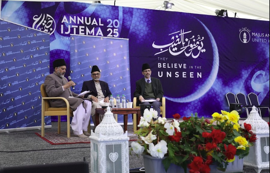Three speakers seated on the Ijtema stage during a session, one of them talking into a microphone, beneath the Annual Ijtema banner
